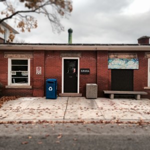 A photo of our office at 24 Trent Lane. A squat red brick building with two windows, a disused metal chimney, with a bunch and two bins (garbage and recycling) outside. It's fall in the photo, so there are some leaves scattered on the ground from nearby trees.